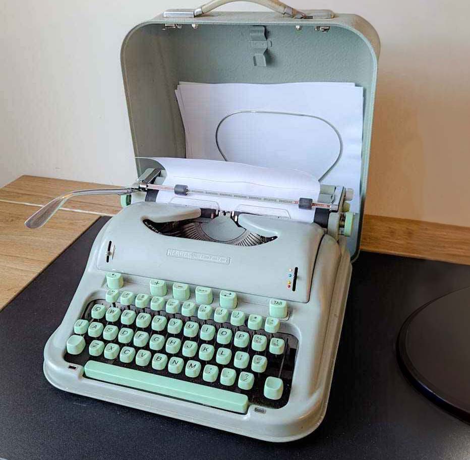 Photograph of a vintage mint-green Hermes 3000 typewriter, open in its travel case with a blank sheet of paper loaded
